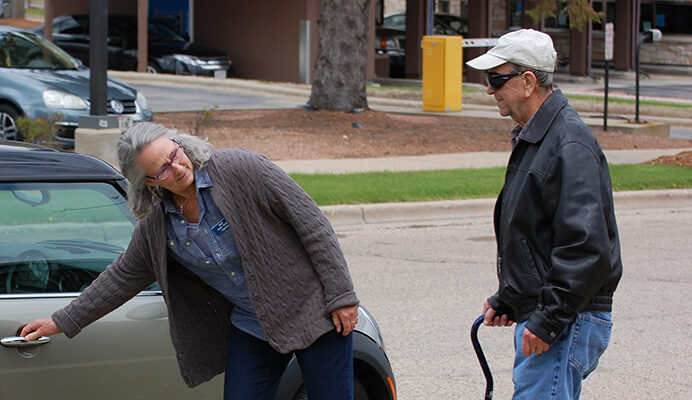 A woman opening the passengers side door of a car for an elderly man with a cane