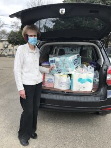 A woman standing in front of an station wagon with its trunk open revealing donated toilet paper and other items