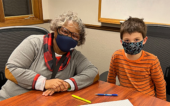 An elderly woman sitting at a desk with a young boy with pens and paper in front of them