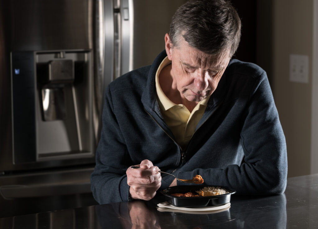 Lonely and depressed senior male sitting alone at kitchen table eating a microwaved ready meal of curry from plastic tray